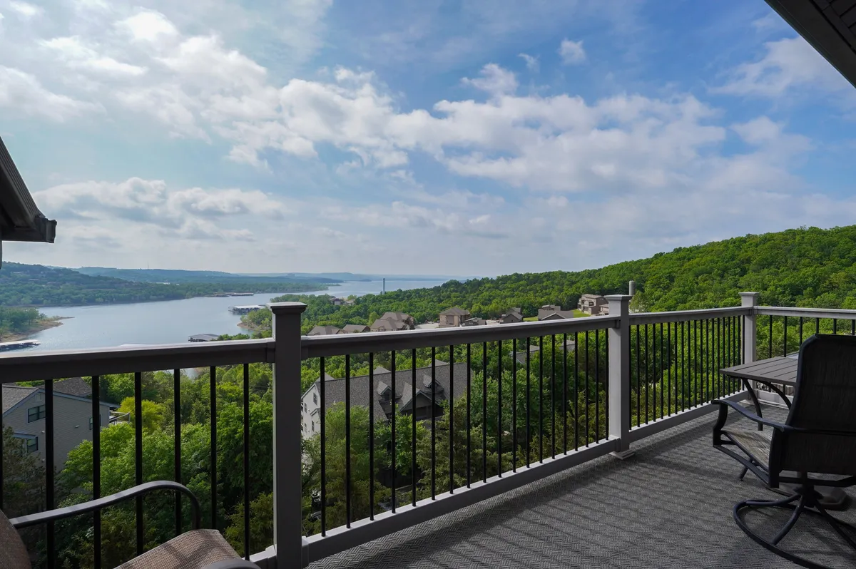 The photo presents a peaceful balcony scene with outdoor furniture, a clear view of the lake, and green hills in the distance. Sunlight gently illuminates the deck, enhancing the serene atmosphere, perfect for morning coffee or unwinding at sunset.