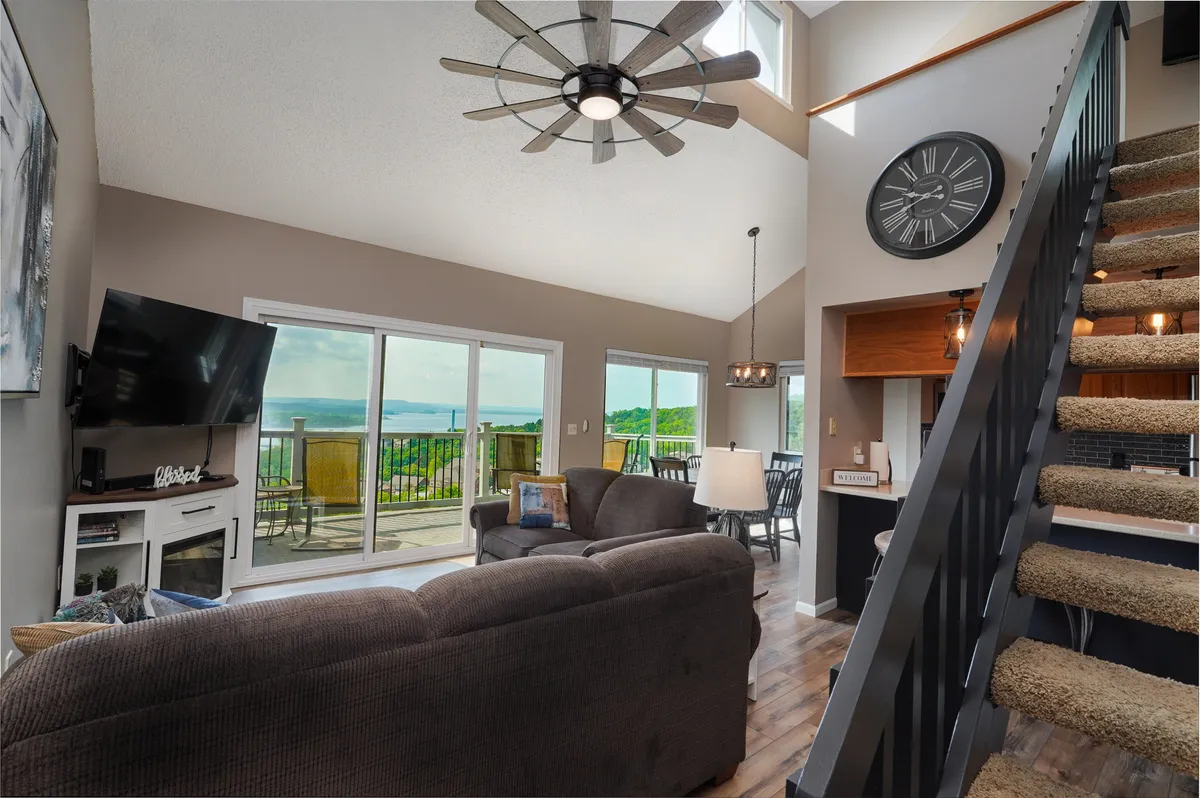 A spacious living and dining area with large windows framing stunning lake views. A comfortable sofa and dining set are arranged to take full advantage of the natural light and scenery. The ceiling fan and tasteful decor add to the room's inviting feel.