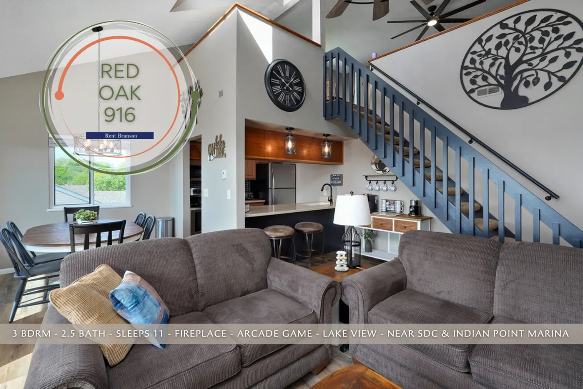 A cozy, open-concept living area featuring comfortable sofas, a visible dining area, and a staircase. Decorative wall art and a fireplace create a welcoming atmosphere. Large windows offer glimpses of the lake beyond, filling the space with natural light.