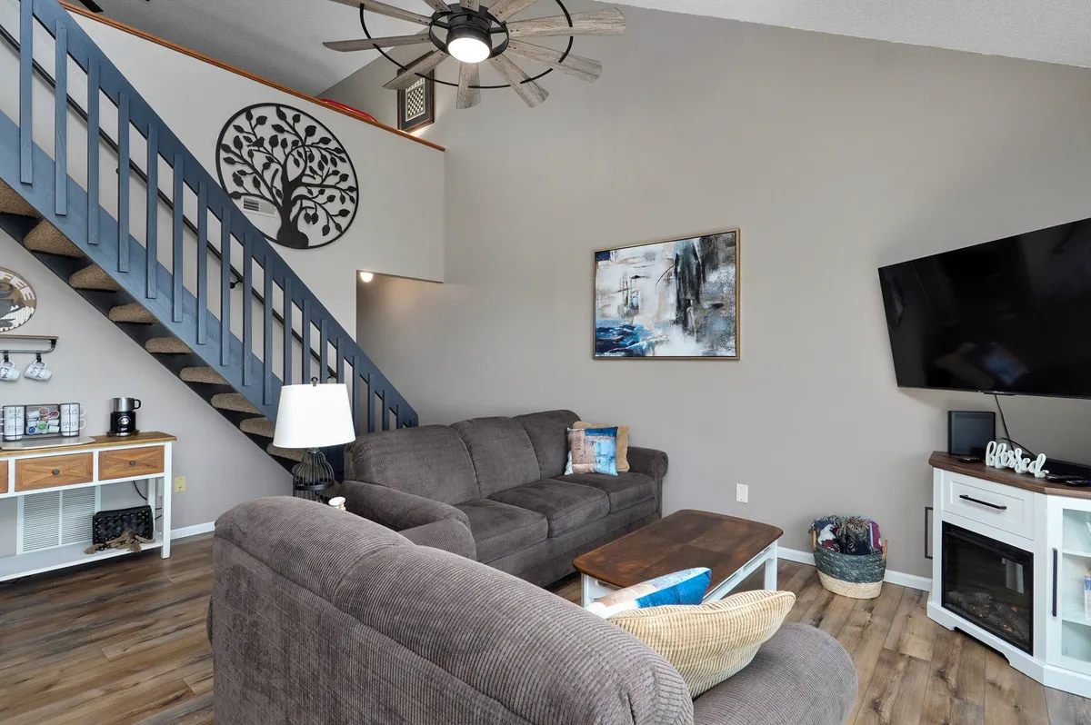 The image showcases a warm, open-concept living area with a stylish sofa, inviting coffee table, modern staircase, tasteful wall art, and a large TV, perfect for relaxing or gathering with family and friends. The space exudes comfort and is ideal for socializing, movie nights, and unwinding after a day by the lake or at nearby attractions.